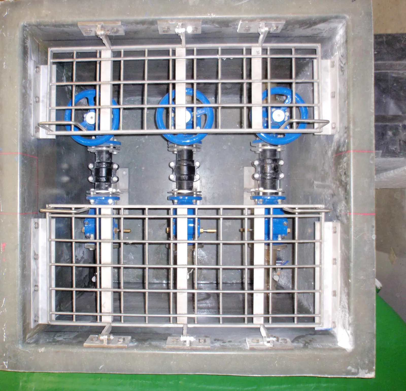 Looking into a Mullaly FRP valve chamber with three blue gate valves behind hinged stainless steel safety grilles